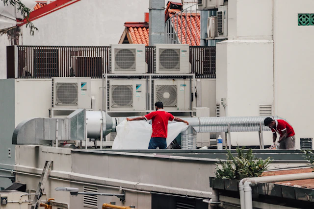 Two workers handling ductwork on a rooftop beside outdoor air conditioning units